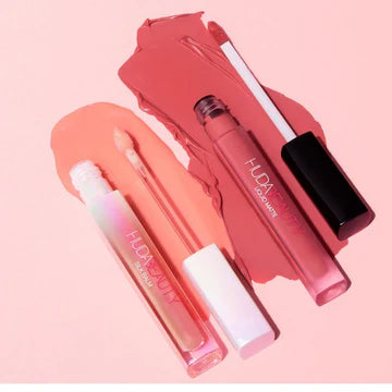 Flatlay of Huda Beauty's Cherry Blossom Lip Set on marble surface – Lucky 04 Liquid Matte Lipstick (coral-pink tube) and Cherry Blossom Silk Balm (gold-cased) beside an open floral pouch with scattered pink petals