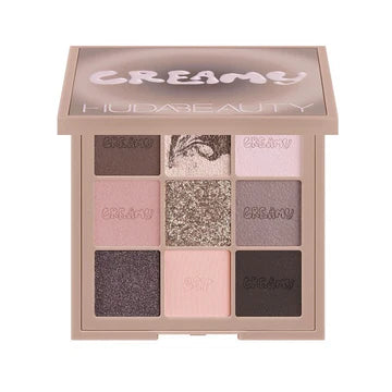 Top-down flatlay of Huda Beauty Creamy Obsessions Greige palette open with all 9 cream-to-powder eyeshadows displayed: grey-brown cream, grey marble, light pink, pinky-grey, grey metallic, blue-grey, purple-grey metallic, neutral pink matte, and black-grey cream