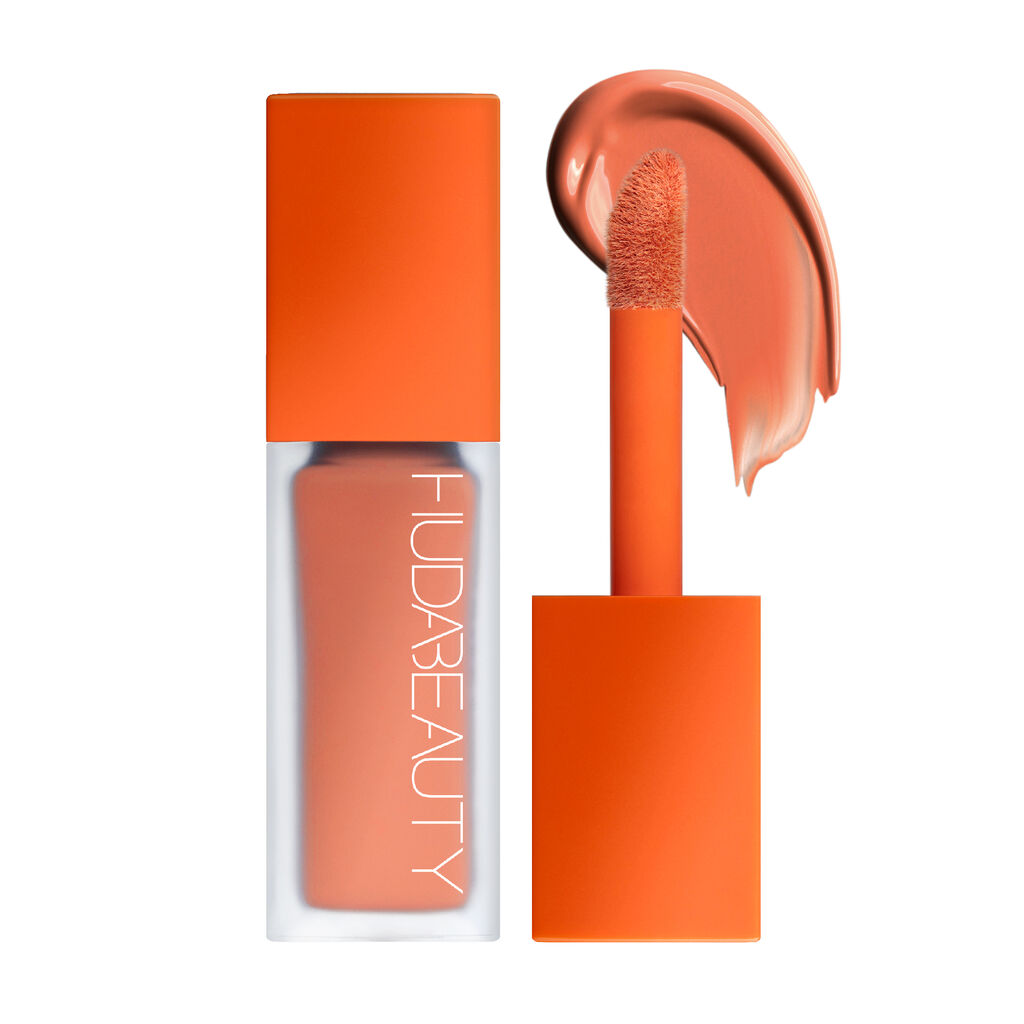 A close-up image of a Huda Beauty #FauxFilter Color Corrector in the shade Mango. The sleek packaging features a frosted tube with a bright orange cap, and the product is shown with its doe-foot applicator and a smooth swatch of the warm orange-toned corrector.