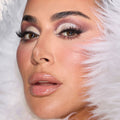 Close-up of a model wearing glamorous icy nude makeup with shimmery silver and mauve eyeshadow, voluminous lashes, glossy nude lips, and soft pink blush, framed by white faux fur.
