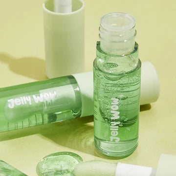 Close-up of SHEGLAM's hydrating lip oil in 'Green Apple Envy,' a glossy green formula designed to moisturize lips. The text 'BIGIRY WARM LIGHTWORK' hints at its lightweight, high-shine finish, housed in a sleek transparent tube