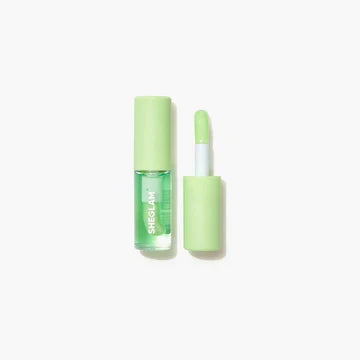 Close-up of SHEGLAM's hydrating lip oil in 'Green Apple Envy,' a glossy green formula designed to moisturize lips. The text 'BIGIRY WARM LIGHTWORK' hints at its lightweight, high-shine finish, housed in a sleek transparent tube