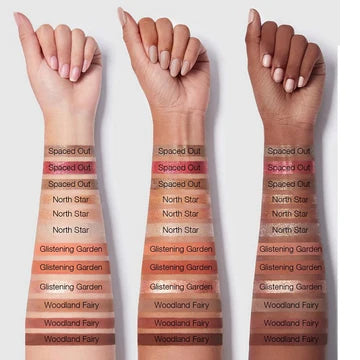 Arm swatches of SHEGLAM 'Spaced Out' liquid eyeshadow trio on light, medium, and deep skin tones. The metallic shades (silver, taupe, and charcoal) show high-impact shimmer on fair skin, buildable iridescence on medium tones, and rich, luminous contrast on deeper complexions. Each shade maintains its shift without ashy undertones